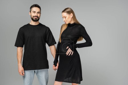 Smiling bearded man in t-shirt and jeans holding hand of stylish and pregnant woman in black dress and standing isolated on grey, concept of expecting parents, husband and wife 