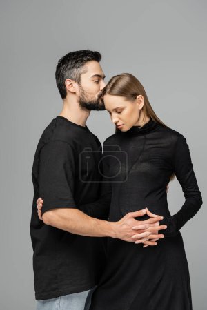 Bearded husband in t-shirt kissing and touching belly of stylish pregnant wife in black dress and standing isolated on grey, concept of expecting parents, happy family 