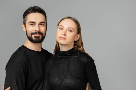 Portrait of smiling and bearded man in black t-shirt looking at camera near stylish fair haired wife and standing isolated on grey, new beginnings and parenting concept 