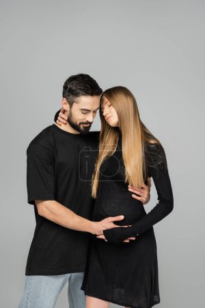 Bearded man in t-shirt and jeans touching belly of stylish and relaxed pregnant wife in dress isolated on grey, new beginnings and expecting parents concept 