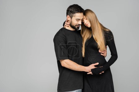 Der bärtige Ehemann im T-Shirt berührt den Bauch der eleganten und blonden schwangeren Frau im Kleid und steht isoliert auf grau, Neuanfang und Erziehungskonzept, Zweisamkeit 
