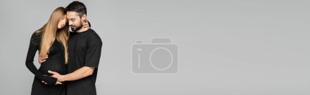 Foto de Marido barbudo en camiseta negra tocando el vientre de la mujer de pelo justo y embarazada de moda, mientras que de pie aislado en gris, nuevos comienzos y el concepto de crianza, bandera - Imagen libre de derechos