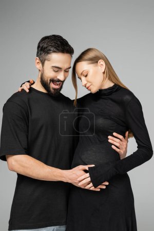 Aufgeregter bärtiger Mann im T-Shirt, der den Bauch berührt und trendige schwangere Frau im Kleid ansieht, während er isoliert auf grauen, neuen Anfängen und Erziehungskonzept steht 
