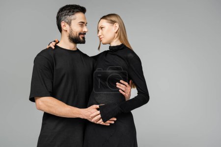 Smiling and bearded husband in black t-shirt hugging and looking at stylish pregnant wife in dress while standing together isolated on grey, new beginnings and parenting concept 