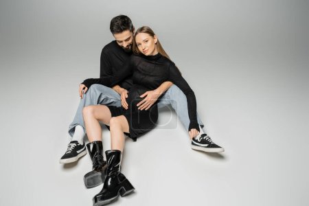 Bearded man in jeans and black t-shirt hugging fashionable pregnant wife in dress and boots while sitting together on grey background, new beginnings and parenting concept 