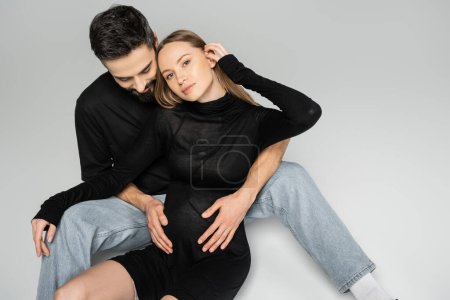 Esposo en camiseta negra y jeans tocando el vientre de la esposa embarazada de moda en vestido mirando a la cámara mientras está sentado en un fondo gris, nuevos comienzos y concepto de crianza 