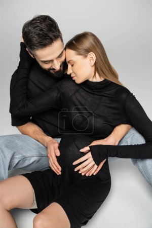 Mujer de pelo justo de moda y embarazada en vestido negro abrazando al marido barbudo mientras están sentados juntos sobre un fondo gris, nuevos comienzos y concepto de crianza 