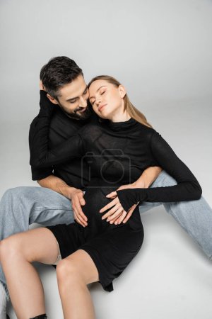 Lächelnder und bärtiger Mann in T-Shirt und Jeans umarmt modische und schwangere Frau, während er zusammen auf grauem Hintergrund sitzt, Neuanfang und Erziehungskonzept 