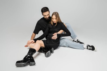 Bearded man in t-shirt and jeans touching belly of fashionable pregnant wife in black dress and boots while sitting on grey background, new beginnings and parenting concept 