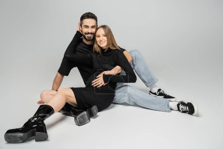 Cheerful fashionable and pregnant woman in black dress and boots looking at camera and hugging husband while sitting on grey background, new beginnings and parenting concept 