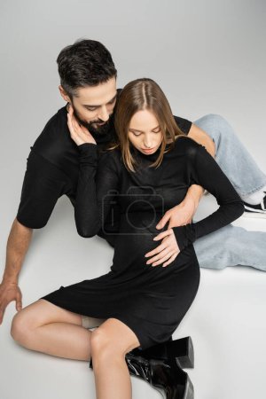 Vista de ángulo alto de la mujer embarazada de moda en vestido negro tocando al marido y mirando el vientre mientras está sentado sobre un fondo gris, nuevos comienzos y concepto de crianza 