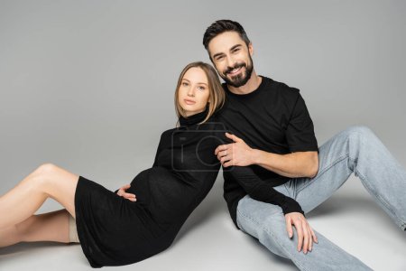 Cheerful husband in t-shirt and jeans looking at camera and hugging trendy pregnant woman in dress while sitting on grey background, new beginnings and parenting concept, husband and wife 