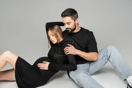Bärtiger Mann in Jeans und schwarzem T-Shirt umarmt stilvolle und schwangere Frau in schwarzem Kleid, während er auf grauem Hintergrund sitzt, Neuanfang und Erziehungskonzept, Mann und Frau 