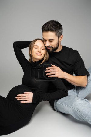 Pleased and bearded man in t-shirt and jeans hugging smiling and pregnant woman while sitting together on grey background, new beginnings and parenting concept 