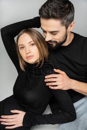 Portrait of bearded husband in t-shirt and jeans hugging stylish and pregnant woman in black dress while sitting on grey background, new beginnings and parenting concept, husband and wife  