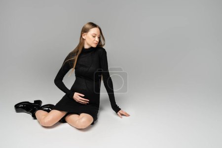 Modische hellhaarige schwangere Frau in schwarzem Kleid und Stiefeln, die wegschaut, während sie auf grauem Hintergrund mit Kopierraum, Neuanfängen und Umstandskonzept sitzt, werdende Mutter 