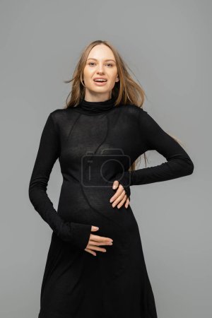Emocionada mujer de pelo pelo y embarazada en vestido negro elegante mirando a la cámara mientras está de pie aislado en gris, nuevos comienzos y el concepto de maternidad, madre-a-ser