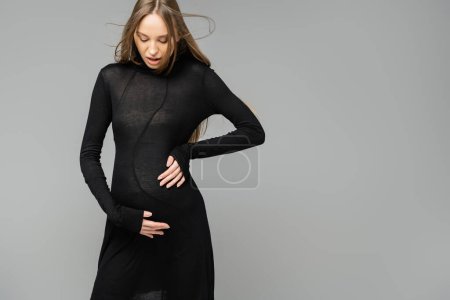 Excited and fashionable pregnant woman in black dress touching belly and looking down while standing isolated on grey, new beginnings and maternity concept, mom-to-be