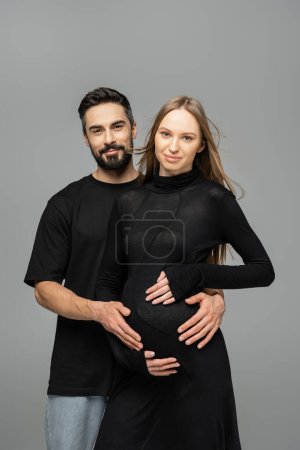 Lächelnder und bärtiger Mann in Jeans und schwarzem T-Shirt blickt in die Kamera und umarmt stilvolle und schwangere Frau und steht isoliert auf grauem, Neuanfang und Erwartungskonzept  