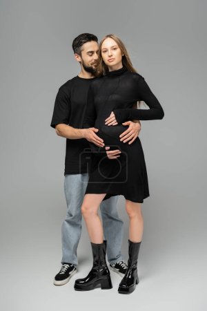 Longitud completa de hombre alegre y barbudo abrazando a la esposa de moda y embarazada mientras está de pie y posando sobre un fondo gris, nuevos comienzos y concepto de anticipación  