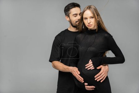 Bearded man in black t-shirt touching belly of fashionable and pregnant wife in dress looking at camera and standing together isolated on grey, new beginnings and anticipation concept  