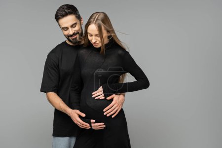 Positive bearded man hugging and looking at belly of pregnant wife in stylish and black dress while standing isolated on grey, new beginnings and anticipation concept  