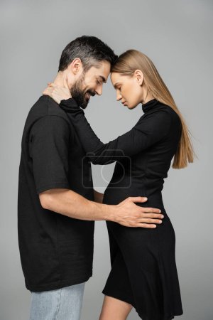 Photo for Side view of stylish and pregnant fair haired woman in black dress hugging smiling and bearded husband while standing with closed eyes isolated on grey, new beginnings and anticipation concept - Royalty Free Image