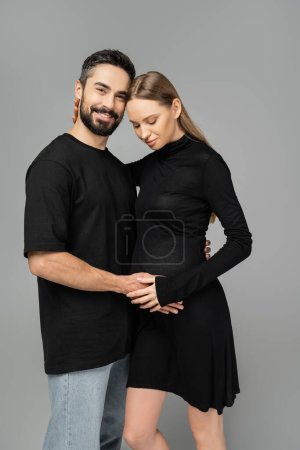 Positiver bärtiger Mann in Jeans und T-Shirt umarmt trendige schwangere Frau in schwarzem Kleid und blickt isoliert in die Kamera auf grau, Neuanfang und Erwartungskonzept  