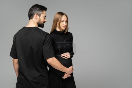 Bearded man in black t-shirt touching belly of trendy and pregnant wife in dress while standing together isolated on grey with copy space, new beginnings and anticipation concept  