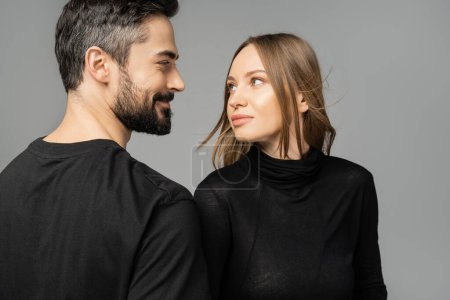 Retrato de mujer elegante de pelo rubio mirando al marido sonriente y barbudo en camiseta mientras están de pie juntos aislados en gris, nuevos comienzos y concepto de anticipación  
