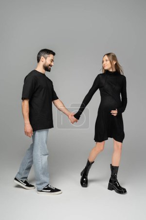 Volle Länge der stilvollen und schwangeren Frau in schwarzem Kleid, die Hand des lächelnden Ehemannes haltend und gemeinsam auf grauem Hintergrund wandelnd, Neuanfang und Erwartungskonzept  