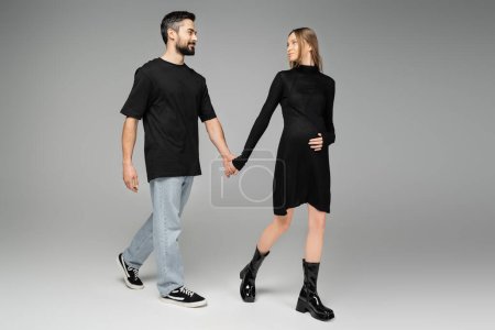 Full length of smiling man in jeans and black t-shirt holding hand of stylish pregnant wife in dress and walking on grey background, new beginnings and anticipation concept, expecting parents 