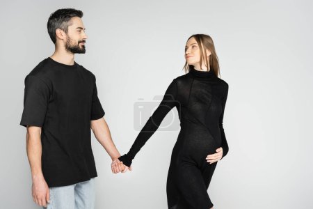 Lächelnder bärtiger Mann im schwarzen T-Shirt an der Hand einer blonden und schwangeren Frau in stilvollem Kleid auf grauem Hintergrund, Neuanfang und Erwartungskonzept, werdende Eltern 