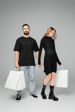 Volle Länge der stilvollen und schwangeren Frau in schwarzem Kleid, die Einkaufstaschen und die Hand ihres Mannes hält, während sie auf grauem Hintergrund steht, Neuanfänge und Elternschaft-Konzept  