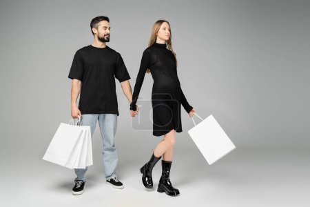 Fashionable pregnant woman in black dress holding shopping bags and hand of husband while walking on grey background, new beginnings and parenthood concept  
