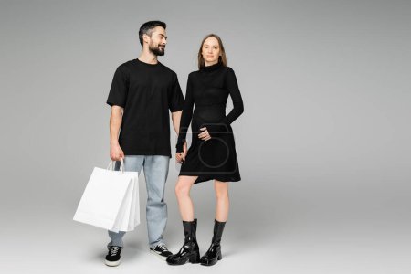 Positive and bearded man holding shopping bags and hand of trendy pregnant wife touching belly and standing together on grey background, new beginnings and shopping concept  