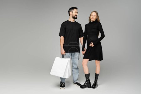 Fröhlicher bärtiger Mann mit Einkaufstüten und Blick auf trendige schwangere Frau im schwarzen Kleid auf grauem Hintergrund, Neuanfang und Elternschaft, Shopping und Erwartungskonzept