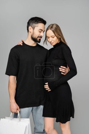 Hombre barbudo en jeans y camiseta sosteniendo bolsas de compras y abrazando a la esposa embarazada con un elegante vestido negro mientras está de pie aislado en gris, nuevos comienzos y el concepto de paternidad  