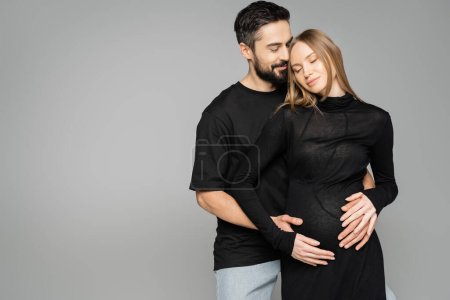 Positiv bärtiger Ehemann im schwarzen T-Shirt umarmt stilvolle schwangere Frau in Kleid und geschlossenen Augen und steht isoliert auf grau, Neuanfang und Erwartungskonzept  
