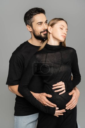 Hombre morena sonriente y barbudo en camiseta negra abrazando a la esposa cabellera y embarazada y mirando hacia otro lado aislado en gris, nuevos comienzos y concepto de anticipación  