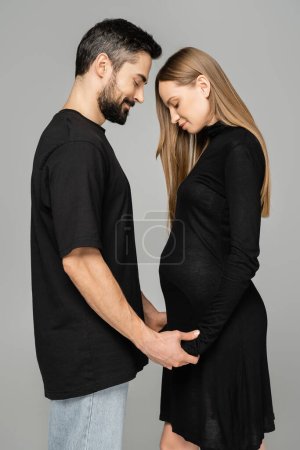 Photo for Side view of bearded man in jeans and t-shirt holding hands of long haired, stylish and pregnant woman while standing isolated on grey, new beginnings and anticipation concept, husband and wife - Royalty Free Image