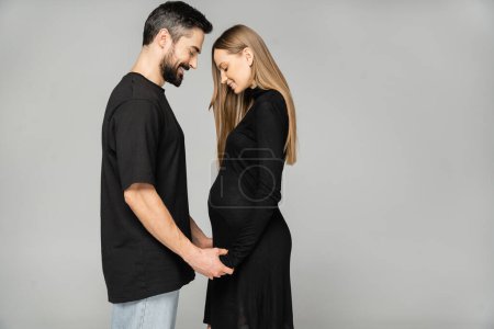 Photo for Side view of positive pregnant and stylish woman in black dress holding hands of bearded man while standing isolated on grey, new beginnings and anticipation concept, husband and wife - Royalty Free Image