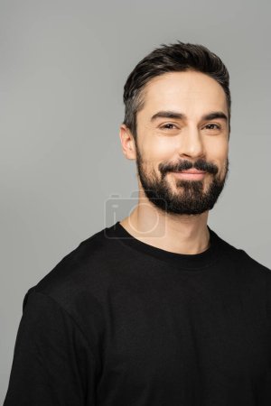 Porträt eines positiven bärtigen und brünetten Mannes im lässigen schwarzen T-Shirt, der isoliert in die Kamera blickt, während er auf einem grauen, maskulinen Schönheitskonzept steht, selbstbewusst und charismatisch 