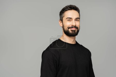 Porträt eines fröhlichen brünetten und bärtigen Mannes im lässigen schwarzen T-Shirt, der in die Kamera lächelt, während er isoliert auf einem grauen, maskulinen Schönheitskonzept steht, selbstbewusst und charismatisch 