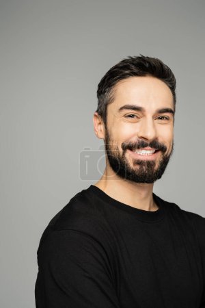 Porträt eines selbstbewussten und fröhlichen bärtigen Mannes im lässigen schwarzen T-Shirt, der in die Kamera blickt und isoliert auf einem grauen, maskulinen Schönheitskonzept steht 