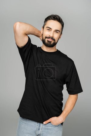 Relaxed and smiling bearded man in black t-shirt and jeans holding hand in pocket and touching neck while looking at camera isolated on grey, masculine beauty concept, confident and charismatic 