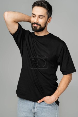 Porträt eines entspannten und bärtigen Mannes in lässigem schwarzem T-Shirt und Jeans, der die Hand in der Tasche hält und wegschaut, während er isoliert auf einem grauen, maskulinen Schönheitskonzept steht 