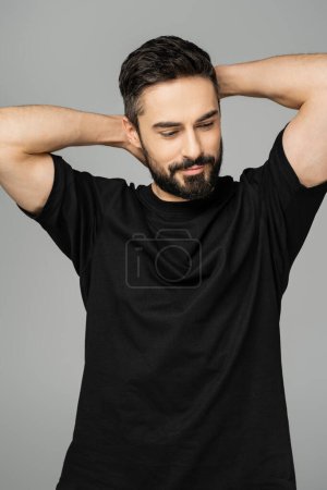 Porträt eines entspannten brünetten und bärtigen Mannes in schwarzem lässigem T-Shirt, das Hals und Kopf berührt, während er wegschaut und isoliert auf einem grauen, männlichen Schönheitskonzept steht 