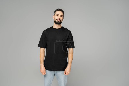 Confident and relaxed bearded and brunette man in black t-shirt and jeans looking at camera while standing isolated on grey, masculine beauty concept, fashion-forward 