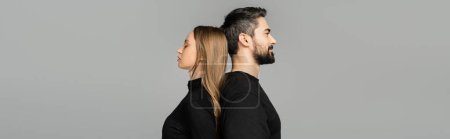 Téléchargez les photos : Vue latérale de l'homme détendu et barbu en t-shirt noir debout dos à dos avec femme aux cheveux blonds isolé sur gris avec espace de copie, concept de relation mari-femme, bannière - en image libre de droit